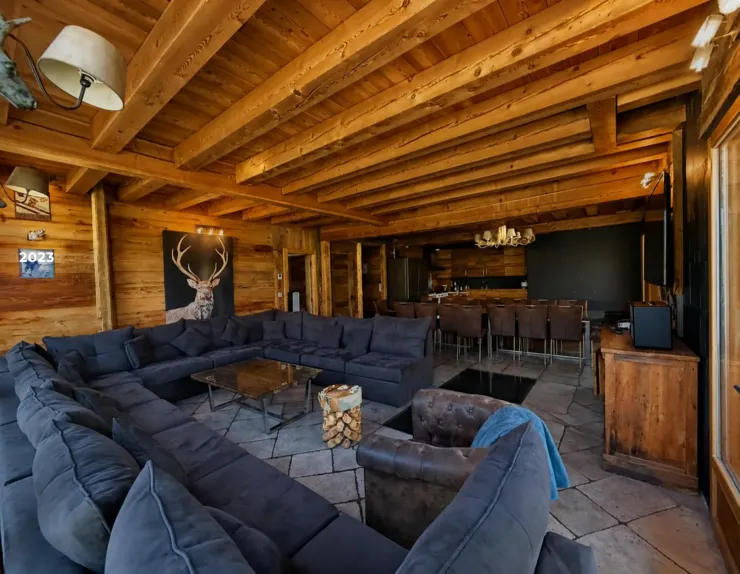 Grand salon du Chalet D à Vars avec canapé d'angle, fauteuil chesterfield, plafond en poutres de bois et vue sur la salle à manger avec sa grande table pour 19 personnes
