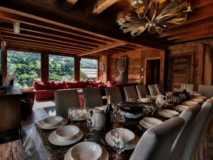 Salle à manger du Chalet D à Vars avec grande table en bois dressée pour 19 convives, appareils à raclette, lustre en bois flotté, plafond à poutres apparentes, ouverte sur le séjour et ses baies vitrées donnant sur la vallée verdoyante